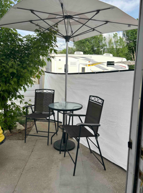 A small outdoor seating area with two black chairs and a table under a large umbrella, surrounded by greenery.